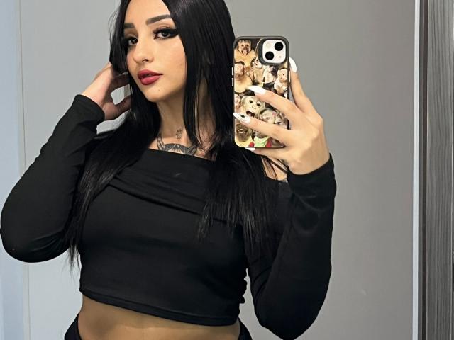 ScarletFeline, com longos cabelos pretos, vestindo uma cropped preta de manga comprida, segurando um celular com um wallpaper de um grupo de K-pop, tem tatuagens no peito e está usando batom vermelho..