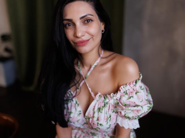 Bellissima Beatrice, con i suoi lunghi capelli neri, sorridente, indossa un abito floreale con una spalla scoperta, mostrando il seno, e ha un sorriso adorabile, mentre è in piedi in una stanza con una luce soffusa..