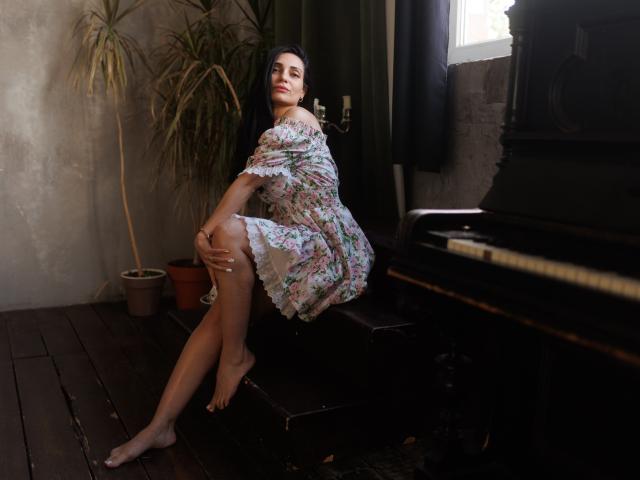 BeaLovely, con lunghi capelli scuri, vestito floreale, piedi nudi, seduta su una panca per pianoforte, mano destra sul ginocchio, gamba sinistra sollevata, pavimento in legno scuro, pianta in vaso, luce proveniente dalla finestra..