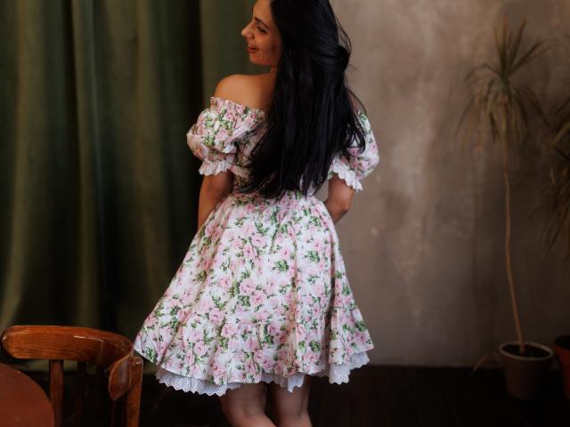 Beatrice, con lunghi capelli neri, indossa un abito rosa con fiori e spalline scoperte, è in piedi con la schiena rivolta verso la fotocamera, mostrando i glutei e un sorriso malizioso, in una stanza accogliente..