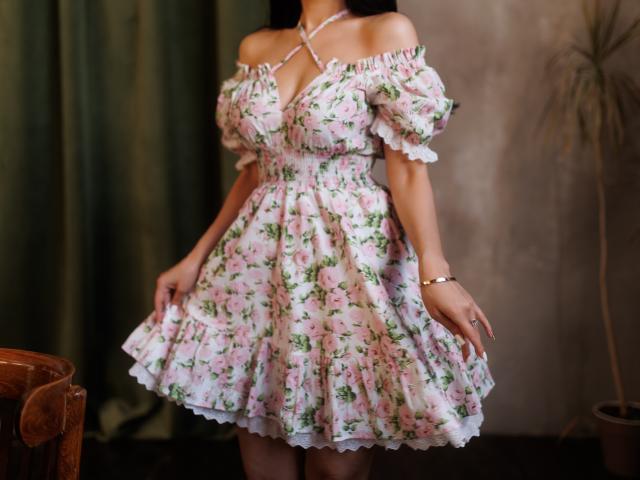 Beatrice, elegante in un abito floreale con rose rosa, a spalle scoperte, con maniche a sbuffo e orlo arricciato, che tiene la gonna, indossa una collana bianca e un braccialetto dorato..