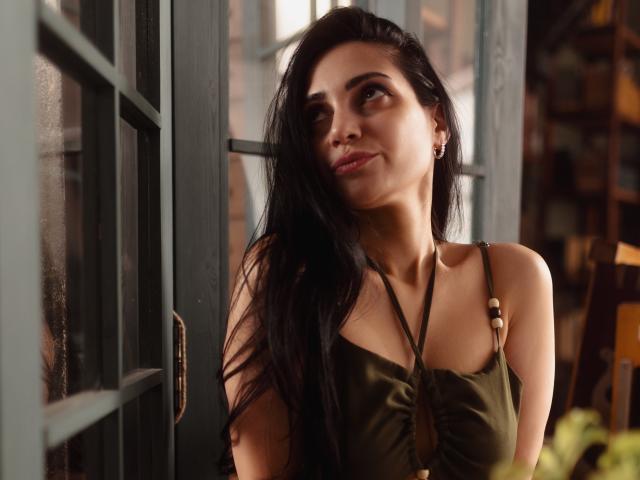 Beatrice, con lunghi capelli neri, un vestito verde e degli orecchini, guarda verso l'alto con un leggero sorriso, in piedi accanto a una finestra con cornice verde, i suoi seni appena visibili sotto il vestito..