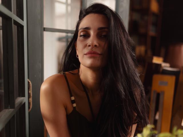 Beatrice, con lunghi capelli neri, occhi marroni e pelle abbronzata, indossa una canotta nera, guarda leggermente verso il basso con un sorriso appena accennato, seduta vicino a una finestra verde..
