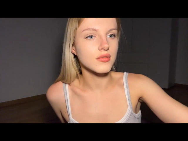 JulissaUrso heeft bleke huid, blond haar en roze lippen. Ze draagt een witte tanktop, waardoor haar kleine borsten zichtbaar zijn. Haar blauwe ogen zijn licht geopend en ze frumelt..