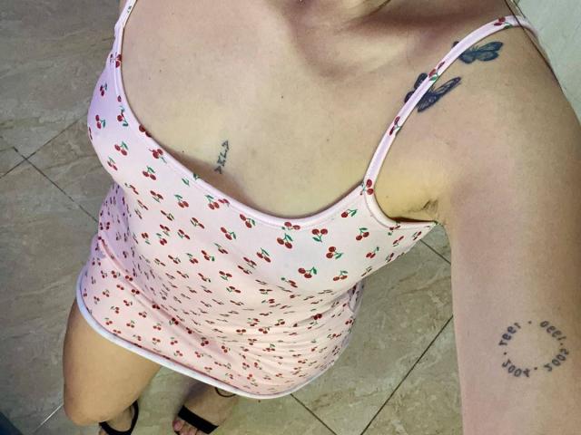 SophiaMarx in a pink cherry-print dress, showing her tits and small ass. She has tattoos on her right shoulder and left arm, wearing black sandals.