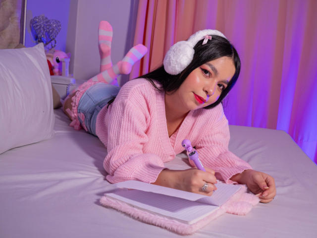 LissyCarter está deitada em uma cama, vestindo pijamas de malha rosa, meias listradas de rosa, fones de ouvido brancos e fofos, e segurando um vibrador roxo, enquanto escreve em um caderno com bordas rosa..