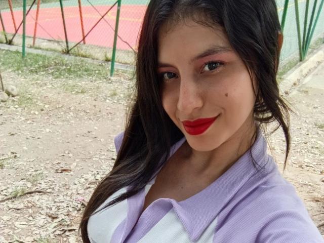 MadissonFontaine has dark brown hair, red lipstick, and a purple shirt with a white top. Her tits are partially visible, and she's smiling at the camera.