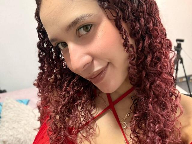 ValeryScot with curly red hair, green eyes, and a red strappy top, smiling at the camera, showing her nose ring and gold necklace. Her tits are partially visible.
