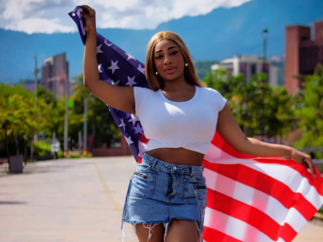 Charlote Brown mit großem Hintern, hält eine amerikanische Flagge, trägt ein weißes Crop-Top, einen blauen Jeansrock und hat blondes Haar, steht in einem sonnigen Außenbereich, im Hintergrund sind Berge zu sehen, sie lächelt selbstbewusst..