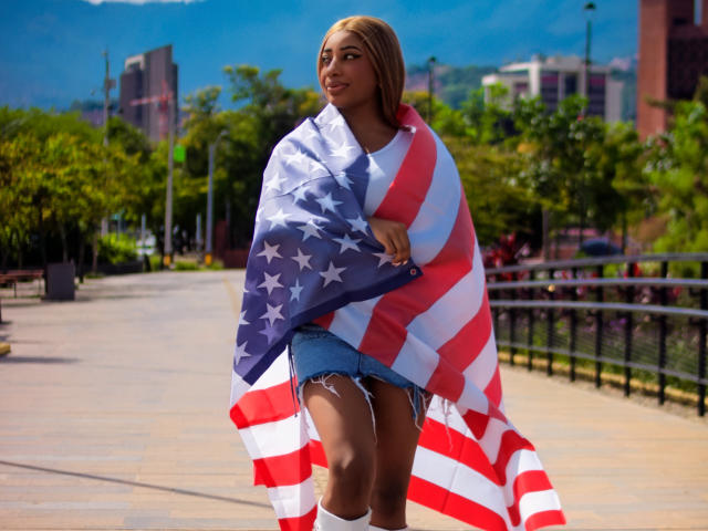 Charlote Brown, con una grande bandiera americana drappeggiata addosso, indossa pantaloncini di jeans e calze bianche, sorride con i capelli castani, in piedi su una banchina assolata, con le montagne sullo sfondo..