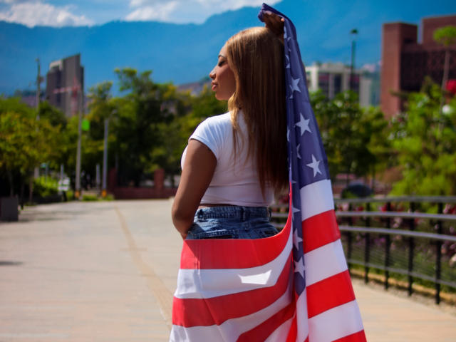 CharloteBrown, avec de longs cheveux bruns, un t-shirt blanc et un short en jean, tenant un drapeau américain, debout sur un trottoir ensoleillé, regardant en arrière avec une expression calme..