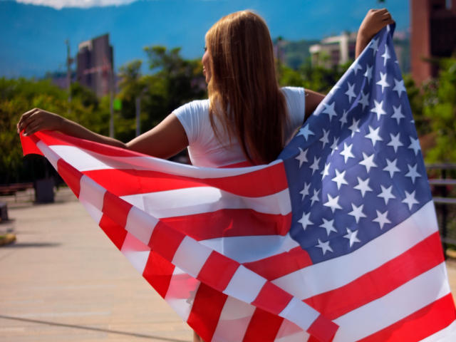 Charlote Brown mit langen blonden Haaren, in einem weißen Hemd, hält eine große amerikanische Flagge, mit dem Rücken zur Kamera, auf einem sonnigen Dach..