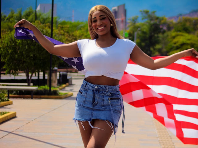 Charlote Brown mit großem Hintern, in einem weißen Crop-Top und einem Jeansrock mit fransigen Säumen, hält amerikanische Flaggen, lächelt, blonde Haare, im Freien, sonnig, blauer Himmel, Berge im Hintergrund..