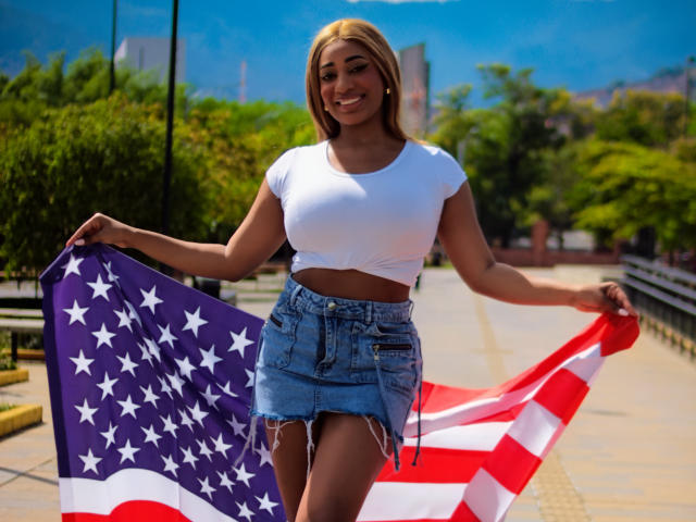 Charlote Brown mit großen Brüsten, lächelnd, hält eine Flagge mit Sternen und Streifen, trägt ein weißes Crop-Top und einen zerrissenen Jeansrock, steht auf einem sonnigen Bürgersteig..