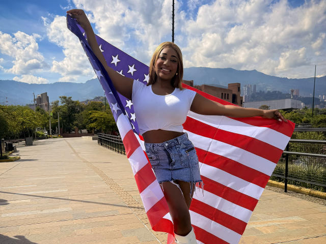 Charlote Brown, mit dunkler Haut, blonden Strähnen, einem weißen Crop-Top, Jeansshorts, hält eine große amerikanische Flagge, lächelt und steht auf einem sonnigen Bürgersteig mit Bergen und blauem Himmel..