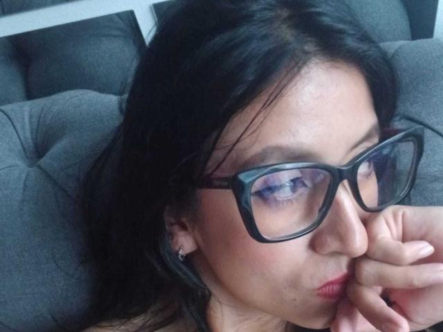 ZarayFeet with black glasses, puffy blue eyes, and pink lips, resting her chin on her hand, showing subtle makeup, and dark wavy hair, on a gray tufted couch.