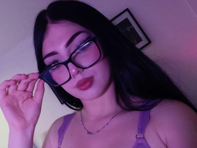 IsabellaaRossi with long black hair, wearing purple glasses, purple bra, and silver necklace, has full red lips and is holding her glasses with a slight pout.