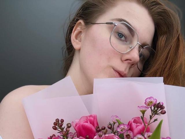 LilliamLechliter, usando óculos, segurando cartões com flores rosa, tem pele clara, cabelo ruivo e um leve sorriso, mostrando seus seios parcialmente escondidos pelos cartões..