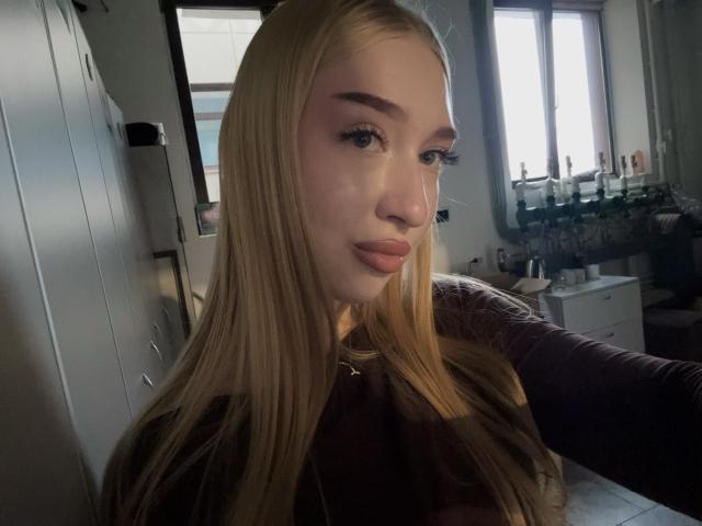 Joette Shigeta, blonde, avec de longs cheveux, du fard à paupières rose, des lèvres pulpeuses et une peau claire, se prend en photo dans une pièce peu éclairée, montrant son visage et sa partie supérieure du corps..