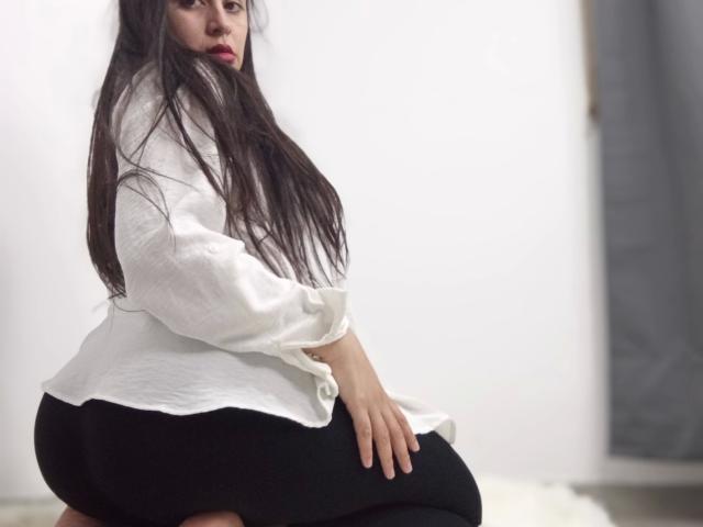 VeliSex - aantrekkelijk met lang donker haar, een wit shirt en zwarte broek, zittend op een kruk, kijkend omhoog met rode lippenstift, haar billen en handen in de voorgrond..