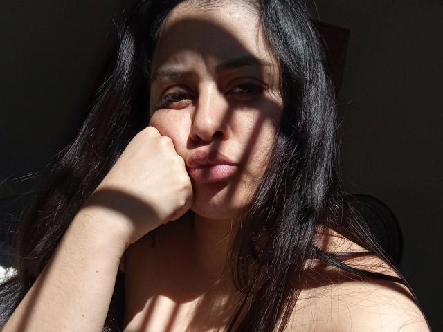 VeliSex, aantrekkelijk met lang zwart haar, een gebruinde huid en volle lippen, leunt haar kin op haar hand, met de zon die haar gezicht in de schaduw legt, waarbij haar blote schouders zichtbaar zijn..