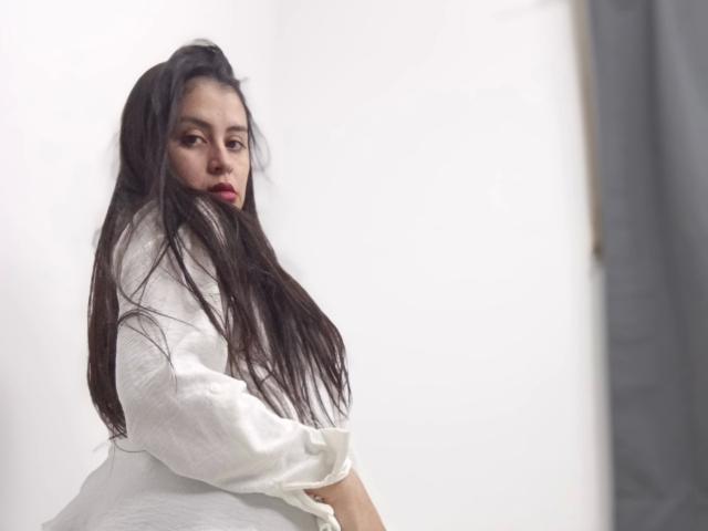 VeliSex - aantrekkelijk met lang zwart haar, een wit shirt en rode lippenstift, kijkt over haar schouder, lichtjes draaiend met armen gekruist, witte achtergrond, zacht licht..