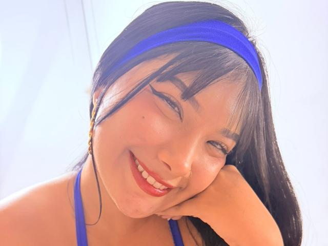 AlejaMiller, con lunghi capelli neri, una fascia blu e rossetto rosso, sorride con gli occhi chiusi, appoggiando la testa sulla mano, indossa un top con spalline blu che mette in mostra il seno..