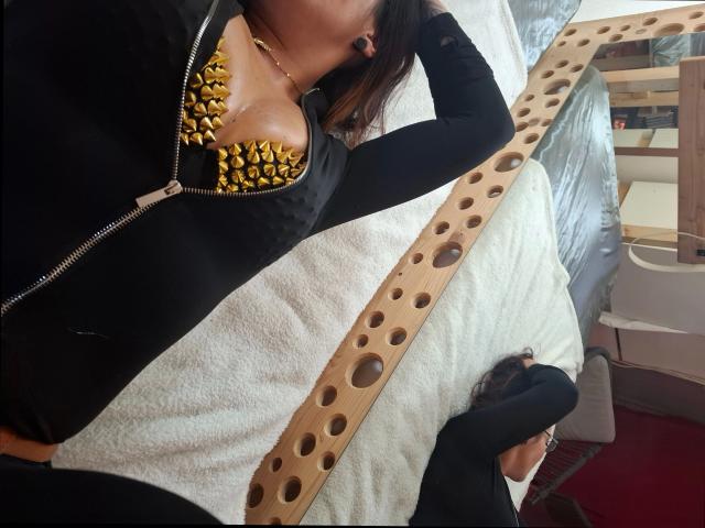 Cinderella-ink-hot with black zip-up jacket, gold-studded black bra showing big tits, black arm, wooden belt with holes, lying on white bed, head down, brown hair, casual.