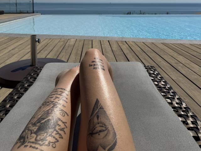 SensualMila's shiny, tattooed legs with intricate designs, one featuring an eye and pyramid, are stretched out on a wicker lounge chair by a blue, infinity pool.