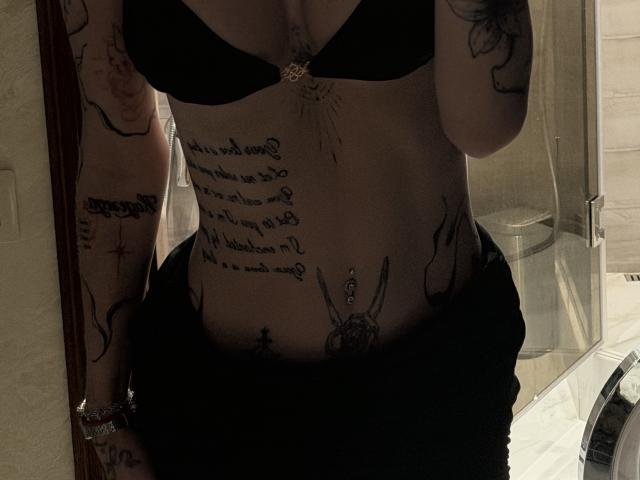 SensualMila met een zwarte bh, tatoeages die haar borst, buik en armen bedekken, een zwarte rok, een zilveren armband, een spiegelbeeld, een zwak verlichte badkamer, zichtbare decolleté en een platte buik..