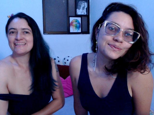 Zwei Frauen mit schwarzem Haar, die dunkle Tanktops tragen und in die Kamera lächeln. Eine von ihnen trägt eine Brille und einen Nasenring, die andere hat kleine Brüste und ein hübsches Gesicht..