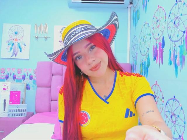 MiaLeroy, avec des cheveux roux, portant une chemise jaune et bleue, un chapeau rayé et du rouge à lèvres rose, souriant à la caméra, montrant légèrement sa poitrine, dans une pièce colorée..