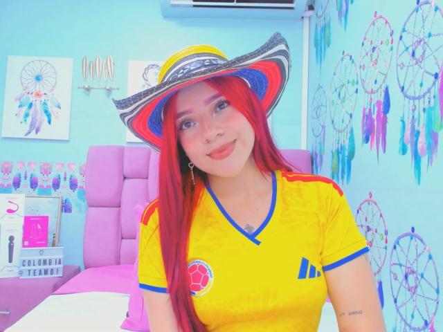 MiaLeroy, avec des cheveux roux, une chemise jaune avec des détails bleus, et un chapeau rouge/violet/blanc, exhibant sa poitrine, assise sur un lit rose, souriant à la caméra..