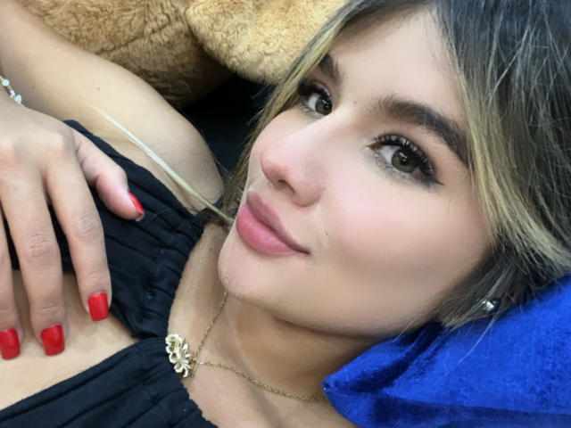 SashaPolet, con cabello rubio, uñas rojas y una blusa negra, acostada, sonriendo, mostrando senos pequeños, un collar dorado, un cojín azul y un osito de peluche marrón en el fondo..