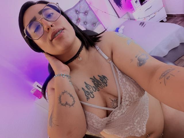 NatalieHarris with glasses, black choker, lace bra, tattoos, black hair, piercing, purple lighting, hand on neck, bracelet, purple walls, bed, and string lights in the background.