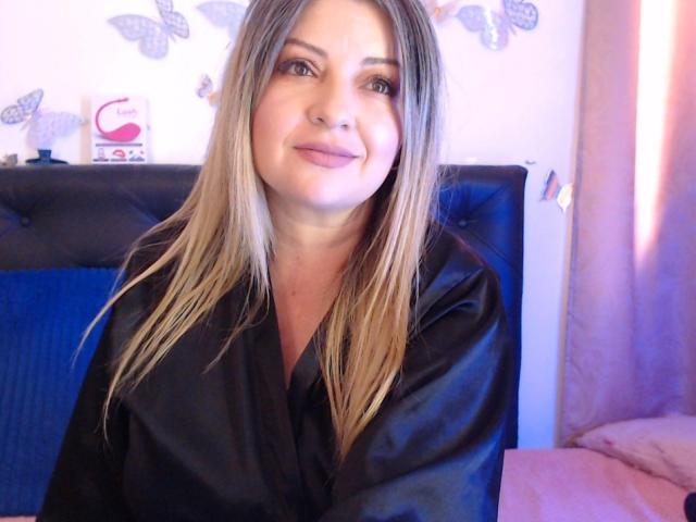 NatalyHami has blonde hair, light skin, and is wearing a black satin robe, sitting with a slight smile. She has medium-sized tits and is looking at the camera.
