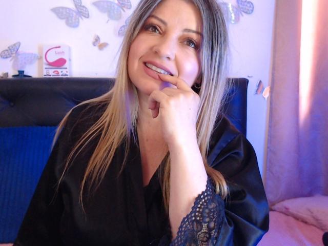 NatalyHami with blonde hair, black silk robe, smiling, biting her finger, showing her cute, small ass, and cute, small tits, in a cozy, pink-lit bedroom.