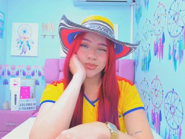 MiaLeroy, avec ses cheveux roux, portant une chemise jaune et bleue, un chapeau de soleil avec des rayures rouges et bleues, reposant la tête sur sa main, souriant doucement. Ses fesses ne sont pas visibles..