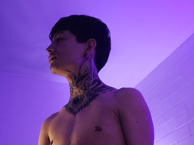 SkylenSnow, torse nu, avec des cheveux foncés, un tatouage au niveau du cou et un piercing au niveau des mamelons, regardant vers la gauche, sous une lumière violette. SkylenSnow a une corpulence moyenne et une légère expression de concentration..