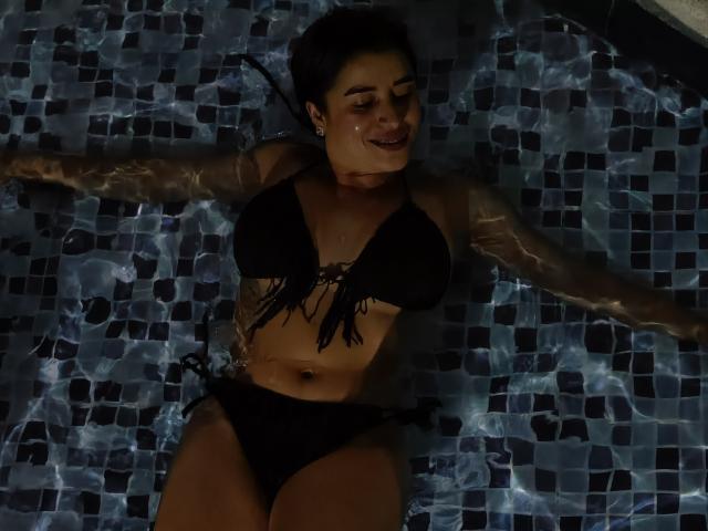 HanaMariana deitada em uma piscina, usando um biquíni preto com parte superior franjada, braços estendidos, água ondulando, cabelo escuro, sorrindo, brincos pequenos, leve bronzeado, seios visíveis, pele escura..