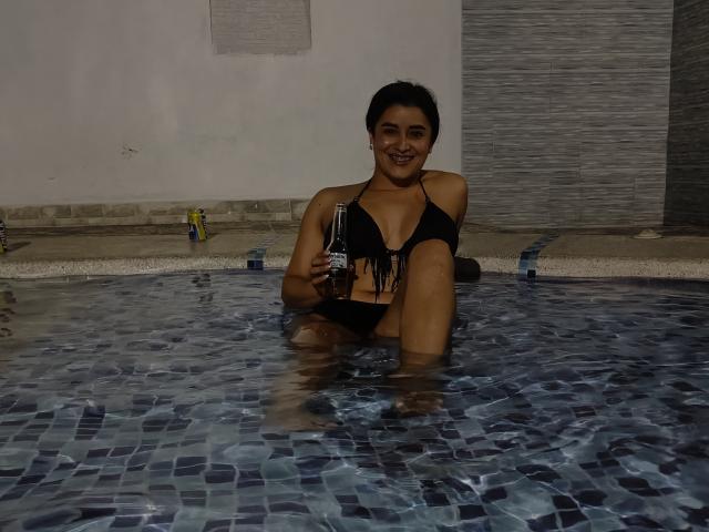 HanaMariana de biquíni preto, sentada em uma piscina com azulejos azuis, segurando uma lata de cerveja, sorrindo, com cabelo escuro e seios médios, em uma piscina interna com pouca luz..