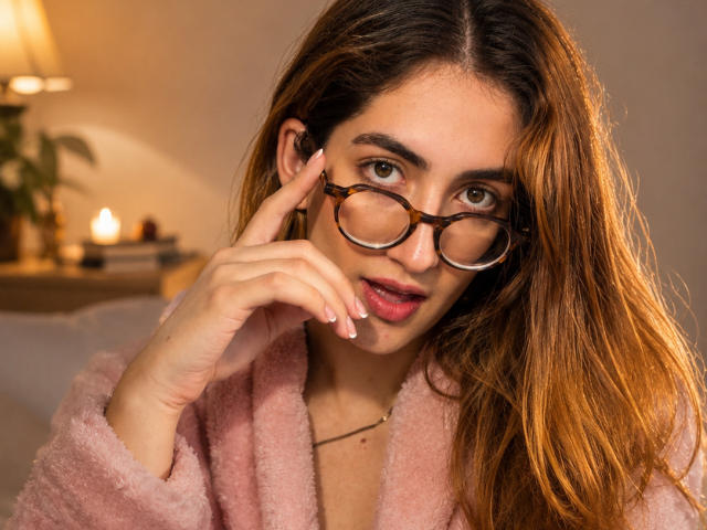 Rousse, avec de longs cheveux bruns, portant des lunettes et une robe rose duveteuse, touchant son nez avec sa main, regardant l'appareil photo avec une expression espiègle et la bouche ouverte..