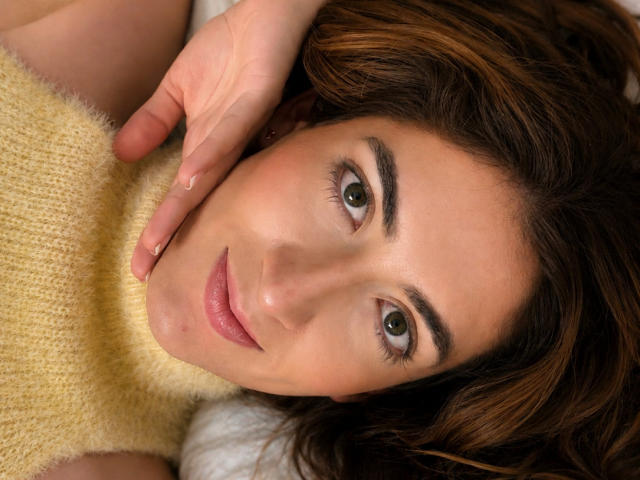 Le visage de RousseauBlossom, allongée, avec des cheveux bruns ondulés, des yeux verts et un sourire discret, portant un pull jaune duveteux, sa main touchant sa joue. Ses seins sont légèrement visibles..