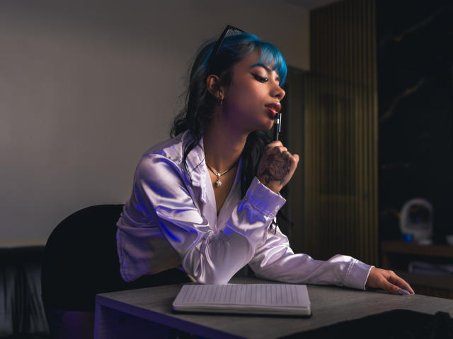 Kazumii, con i capelli blu, la camicia bianca e un dito vicino alla bocca, è seduta a una scrivania con un portatile, appare sexy e sicura di sé, ha dei tatuaggi sulla mano, e la sedia è scura..