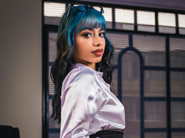 Kazumii, con cabello ondulado azul y negro, vestida con una blusa blanca brillante, mirando por encima del hombro con una ligera sonrisa, sus pequeños senos ligeramente visibles, gafas en la cabeza..