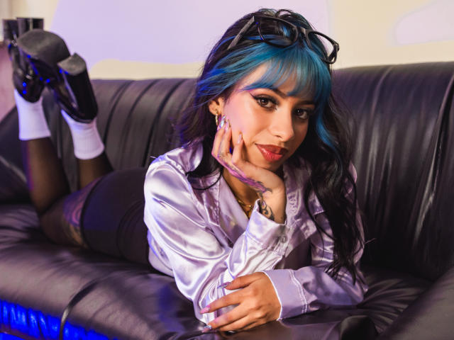 Kazumii, avec ses cheveux bleus et noirs, est allongée sur un canapé en cuir noir, vêtue d'un chemisier blanc brillant, d'une jupe noire et de talons hauts, et elle touche son visage, montrant ses seins..