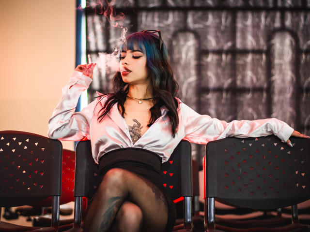 Kazumii, con i capelli blu-neri, una camicia bianca, una gonna nera e seduta su una sedia nera, che fuma, mostra un tatuaggio sul petto, con le gambe incrociate e una collana nera, emanando un'aria sexy e rilassata..