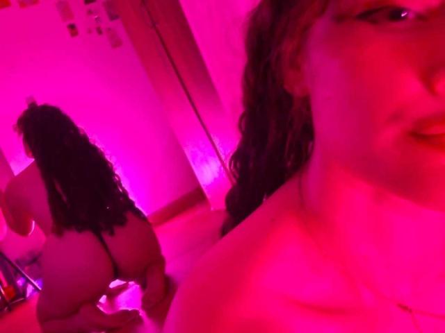DanaStone, com cabelo preto encaracolado, nua, com a nádega voltada para a câmera, em um quarto com iluminação rosa. O rosto de DanaStone em primeiro plano, ligeiramente desfocado. A nádega, as costas e o reflexo de DanaStone visíveis..