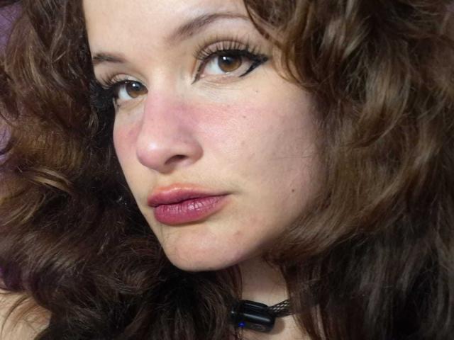 O rosto de DanaStone, com cabelos castanhos cacheados, olhos castanhos, delineador preto, lábios rosados, um leve blush e um colar preto com uma conta azul. Seu nariz é pequeno e sua pele é clara..