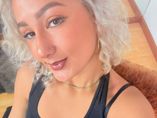 ElenaSaenz with curly white hair, nose ring, and gold chain, wearing black top, looking at camera with glossy pink lips. Her tits are slightly visible under top.